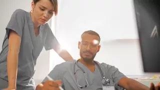 Two healthcare professionals in scrubs make notes as they watch an online learning course at their desk